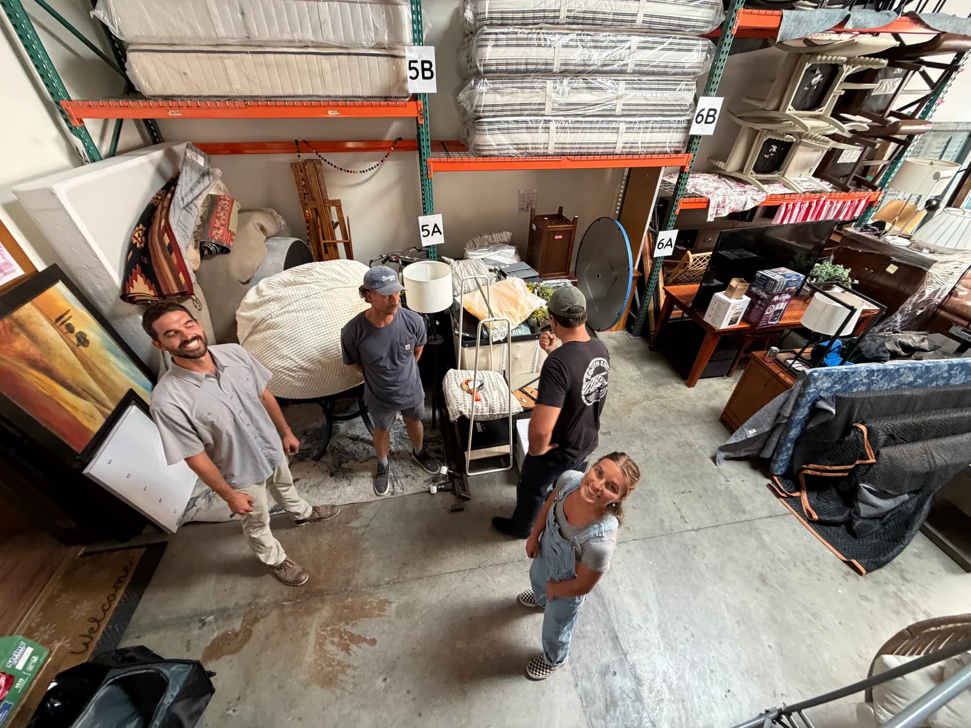 Looking down on four people standing among donated furniture in the Riverbank warehouse, with shrink-wrapped mattresses stacked on the orange racking above them