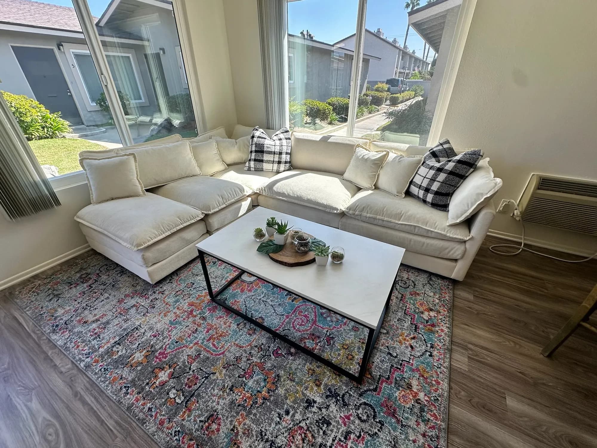 A furnished living room with a cream sectional sofa, a white coffee table, a patterned rug, and small plants, lit by large windows
