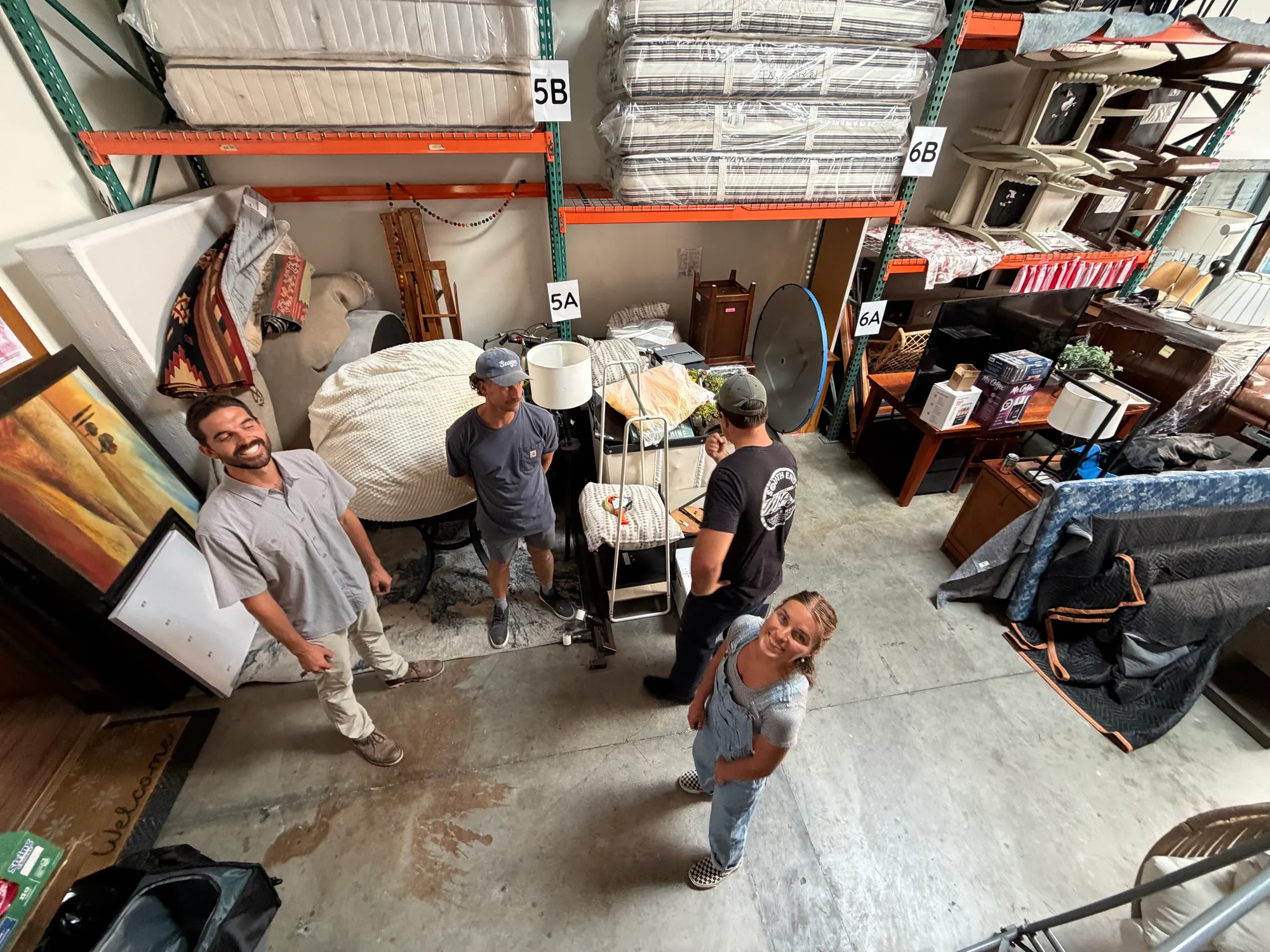 Looking down on four people standing among donated furniture in the Riverbank warehouse, with shrink-wrapped mattresses stacked on the orange racking above them